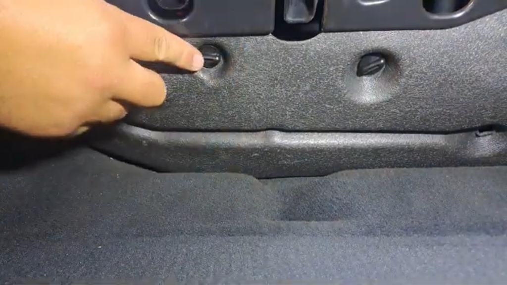 Close-up view of the passenger footwell in an Audi A4 B8, showing a hand pointing to one of the black plastic thumb screws securing the foam trim cover above the cabin filter.